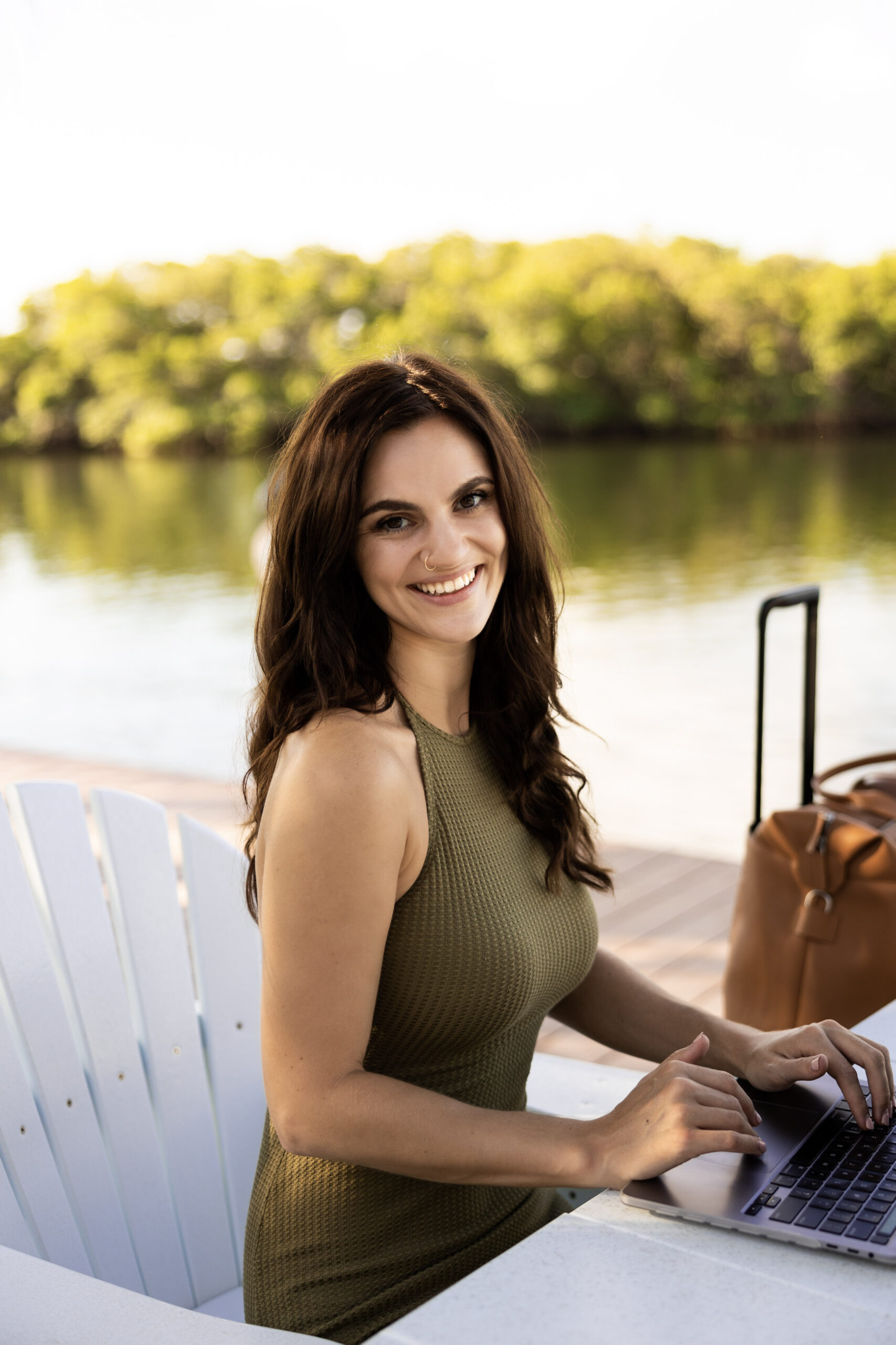 alexandra waggoner sitting at a table by the water on her laptop with her luggage in the background in an aesthetic way